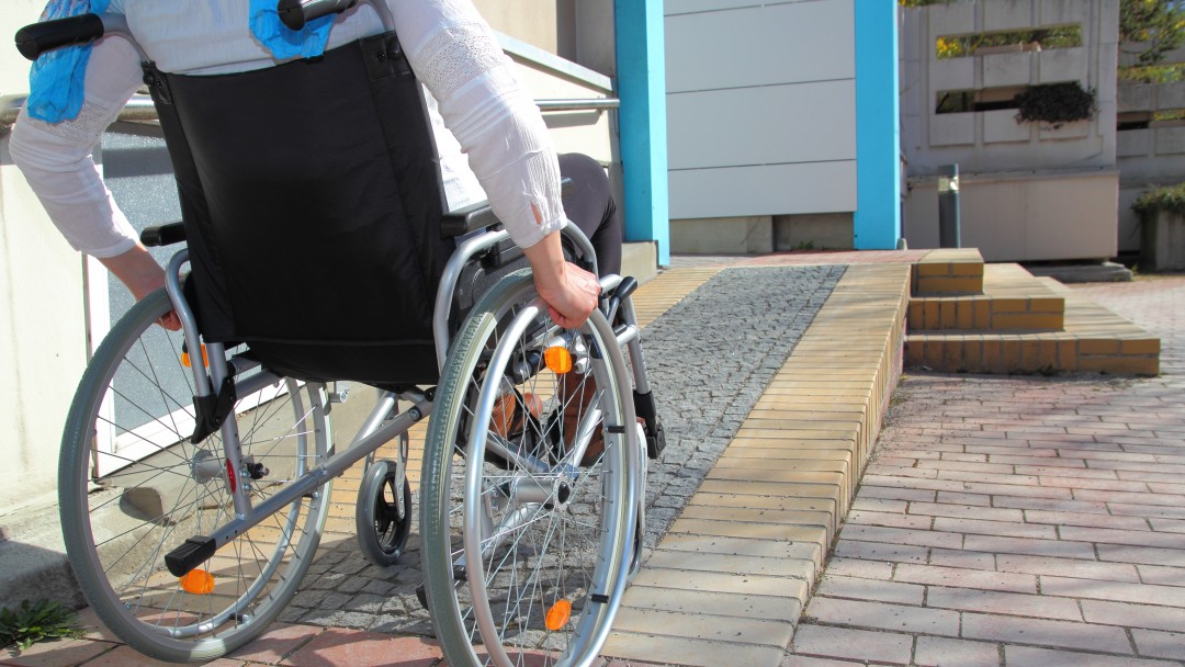 Eine Frau im Rollstuhl befindet sich auf einer Rollstuhlrampe eines Gebäudeeingangs