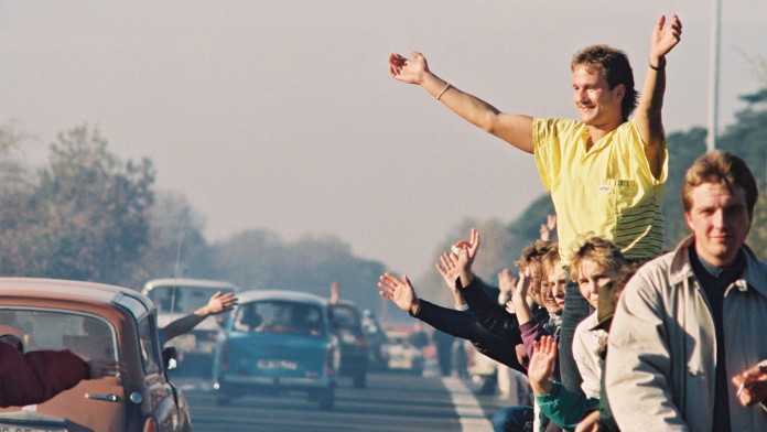 Menschen am Strassenrand winken vorbeifahrenden Autos zu (November 1989)