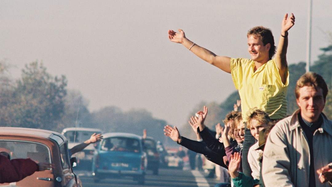 Menschen am Strassenrand winken vorbeifahrenden Autos zu (November 1989)