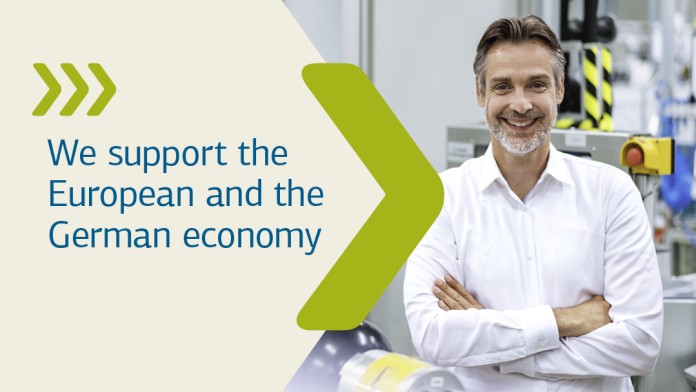 A woman wearing protective clothing and a hard hat stands in front of a wind farm with a laptop