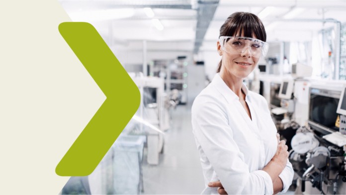 A woman stands in a laboratory with her arms folded and wearing protective goggles