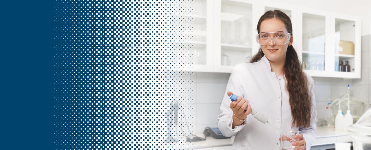A woman in a white coat and protective goggles stands in a laboratory