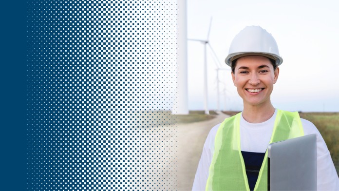 A woman wearing protective clothing and a hard hat stands in front of a wind farm with a laptop