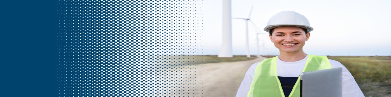 Eine Frau mit Schutzkleidung und Schutzhelm steht mit einem Laptop vor einem Windpark