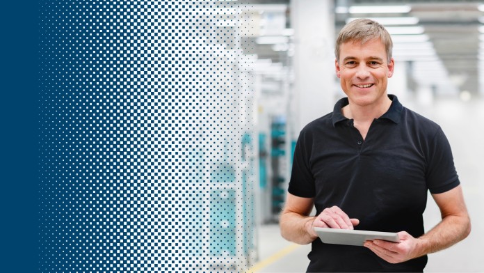 A man with a tablet in his hand stands in a production hall and smiles at the camera