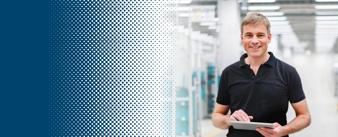 A man with a tablet in his hand stands in a production hall and smiles at the camera