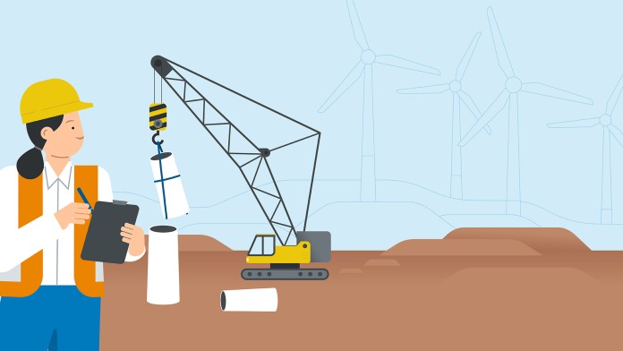 A worker stands with a clipboard in front of a crane erecting a wind turbine