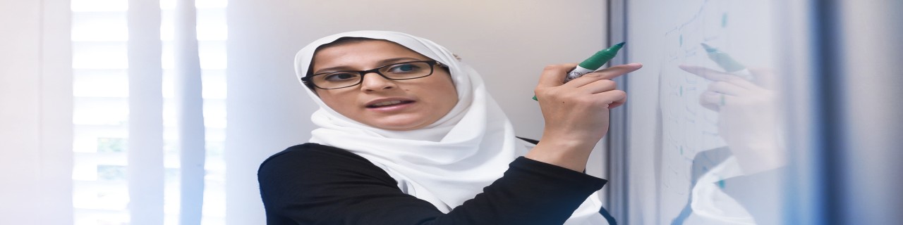 Eine Lehrerin erklärt an einem Whiteboard stehend Inhalte. Es handelt sich um eine Unterrichtssituation. Themen sind Bildung, Wissensvermittlung und Schulentwicklung.