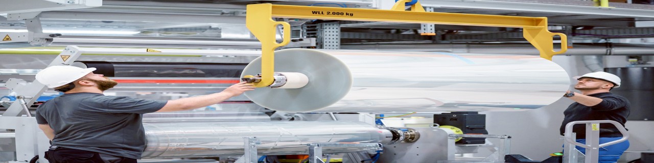 Industriearbeiter montieren eine große Folienrolle in eine(r) Produktionsanlage. Zu sehen ist hier ein Einblick in industrielle Fertigung und Maschinenbetrieb.