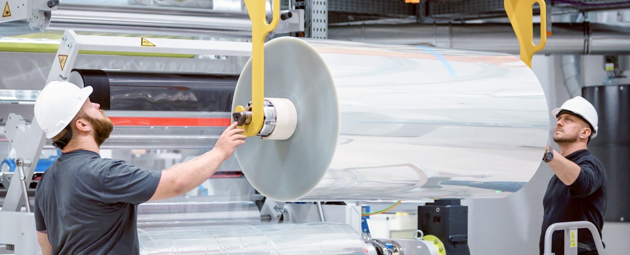 Industriearbeiter montieren eine große Folienrolle in eine(r) Produktionsanlage. Zu sehen ist hier ein Einblick in industrielle Fertigung und Maschinenbetrieb.