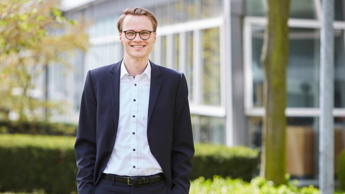 Fabian Peters, Mitarbeiter des Rechtsbereichs der KfW, steht vor einem KfW Gebäude. Er trägt eine Brille und ein weißes Hemd mit dunklem Anzug. Die Hände befinden sich in der Hosentasche.