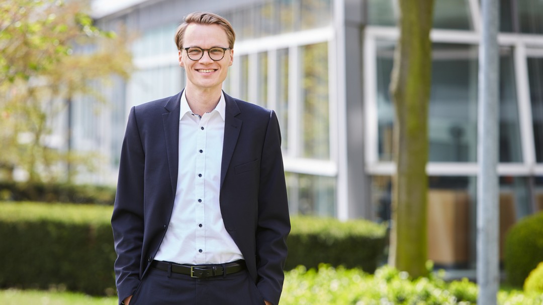 Fabian Peters, Mitarbeiter des Rechtsbereichs der KfW, steht vor einem KfW Gebäude. Er trägt eine Brille und ein weißes Hemd mit dunklem Anzug. Die Hände befinden sich in der Hosentasche.