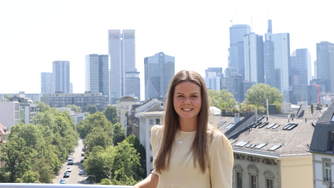 Florine Tüting, Mitarbeiterin des Rechtsbereichs der KfW. Sie trägt ein hellgelbes Oberteil, hinter ihr ist die Skyline von Frankfurt zu sehen.