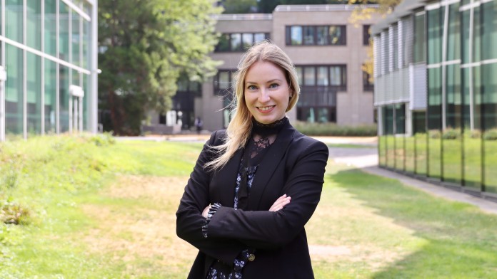 Portraitbild von Jennifer Klein, sie trägt einen schwarzen Blazer und steht mit gekreuzten Armen in einem Innenhof der KfW zwischen zwei Gebäuden. 