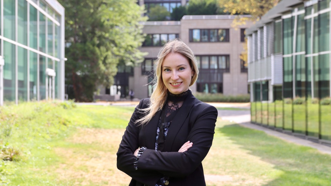 Portraitbild von Jennifer Klein, sie trägt einen schwarzen Blazer und steht mit gekreuzten Armen in einem Innenhof der KfW zwischen zwei Gebäuden. 