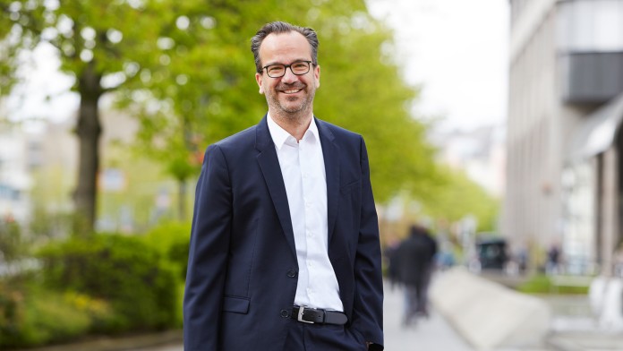 Maximilian Wagner, ein Teamleiter des Rechtsbereichs der KfW, steht vor einem KfW Gebäude. Er trägt eine Brille, ein weißes Hemd und einen dunklen Anzug.