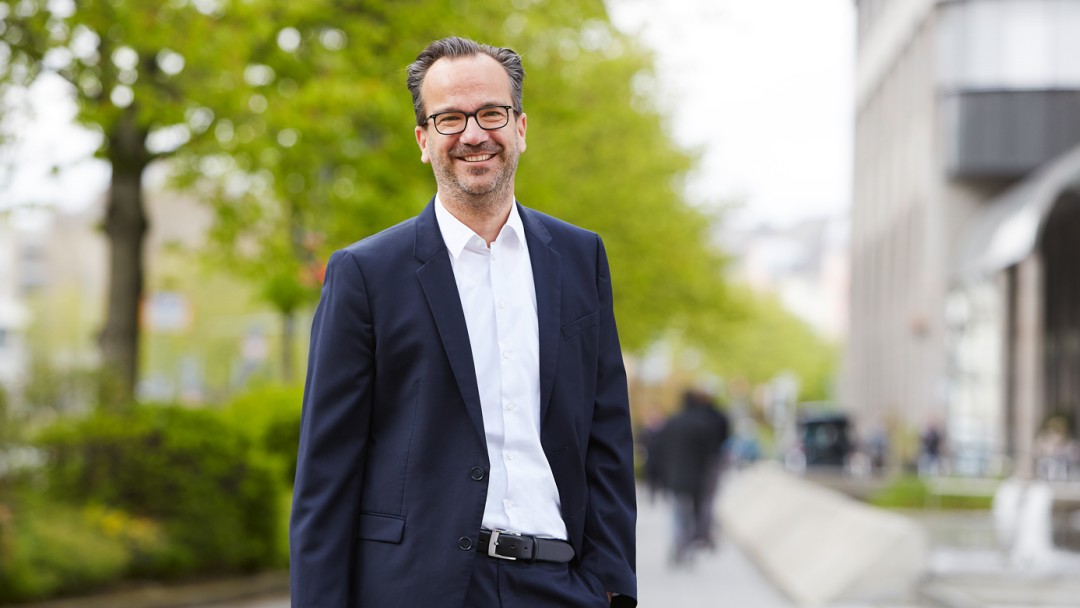 Maximilian Wagner, ein Teamleiter des Rechtsbereichs der KfW, steht vor einem KfW Gebäude. Er trägt eine Brille, ein weißes Hemd und einen dunklen Anzug.