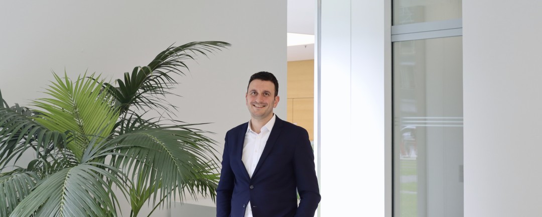 Nick Aghanassir, Mitarbeiter des Rechtsbereichs der KfW steht in einem Foyer der KfW neben einer großen Palmenpflanze. Er trägt ein weißes Hemd mit dunkelblauem Anzug.
