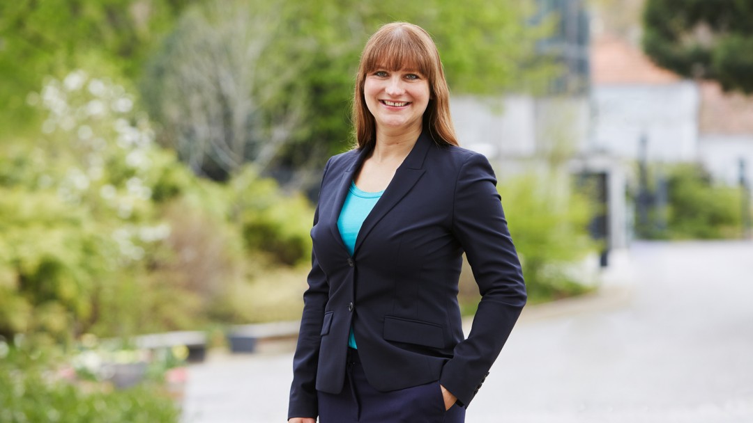 Oriana Corzilius, Mitarbeiterin des Rechtsbereichs der KfW, steht vor einem KfW Gebäude mitgrünen Bäumen. Sie trägt ein türkisfarbenes Oberteil und einen dunklen Hosenanzug.