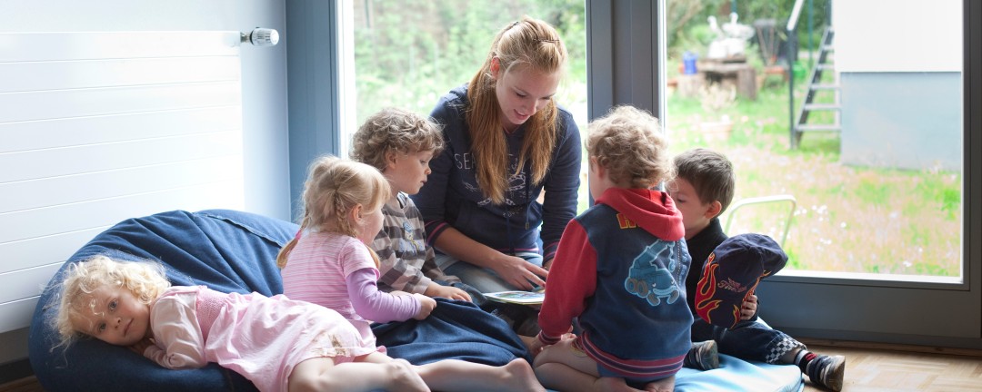 fünf Kinder sitzen um eine Erzieherin herum und hören ihr beim Vorlesen zu