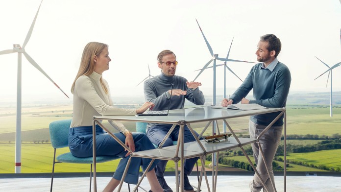 Eine Mitarbeiterin und zwei Mitarbeiter der KfW befinden sich in einer Besprechung, sie sitzen bzw. stehen an einem erhöhten Tisch. Im Hintergrund sind mehrere Windkrafträder auf einem Feld zu sehen.