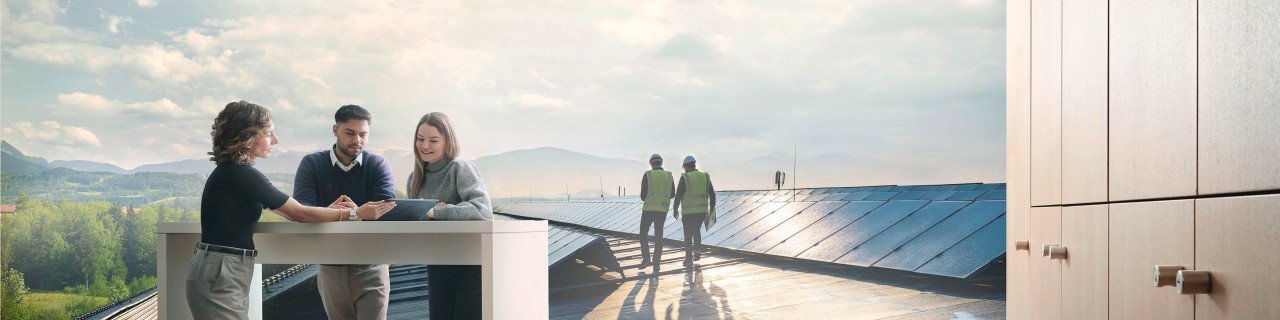 Eine erfahrene Mitarbeiterin der KfW steht mit zwei Auszubildenden an einem Stehtisch und zeigt ihnen ihre nächsten Aufgaben auf einem Tablet. Im Hintergrund sieht man ein Dach mit Solarpanels vor einer Landschaft mit Bergen, zwei Ingenieure mit Warnwesten laufen dort entlang und besprechen sich. 