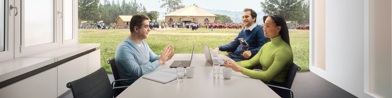 Eine Mitarbeiterin und zwei Mitarbeiter der KfW befinden sitzen zusammen an einem Tisch in einem Besprechungsraum und diskutieren Arbeitsergebnisse. Im Hintergrund ist eine afrikanische Schule zu sehen vor der Schülerinnen und Schüler stehen.