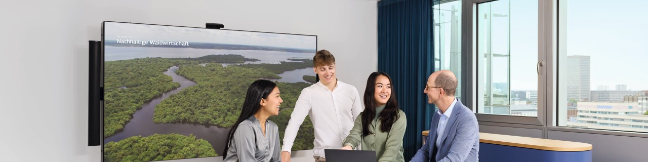 Eine diverse Gruppe (Personen verschiedenen Geschlechts, Alters und Herkunft) sitzt zusammen an einem Besprechungstisch und diskutiert angeregt über nachhaltige Waldwirtschaft. Im Hintergrund ist auf einem großen Monitor ein Bild vom Regenwald zu sehen.