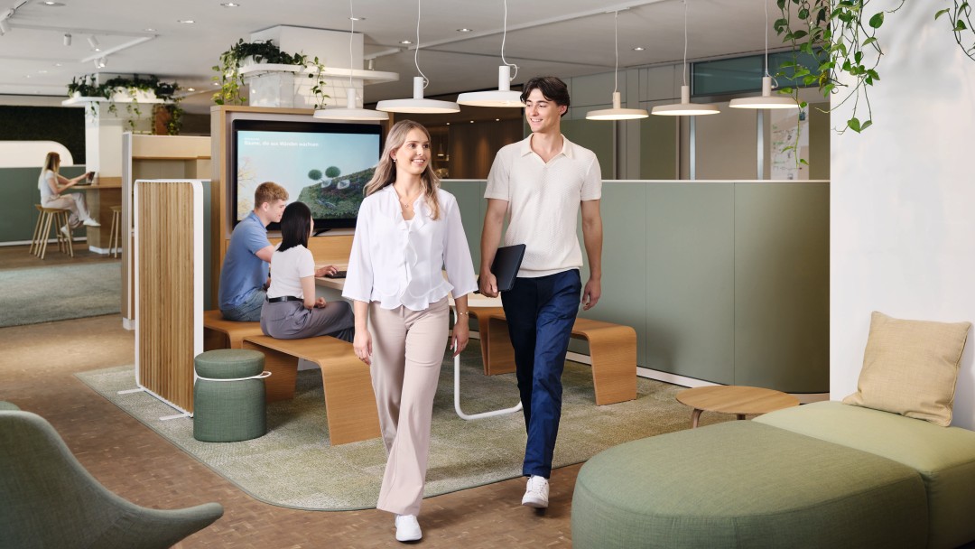 Zwei junge Nachwuchskräfte bewegen sich dynamisch von einer Gruppenarbeit weg, um zu dem nächsten Meeting zu gehen. Sie hat blonde mittellange Haare und trägt eine weiße Bluse sowie beige Hose. Er hat dunkle Haare und trägt ein helles Oberteil sowie dunkelblaue Hose und hält ein Laptop in der Hand. Im Hintergrund sitzen zwei weitere Nachwuchskräfte zusammen, die an dem Thema Verbesserung des Stadtklimas arbeiten.