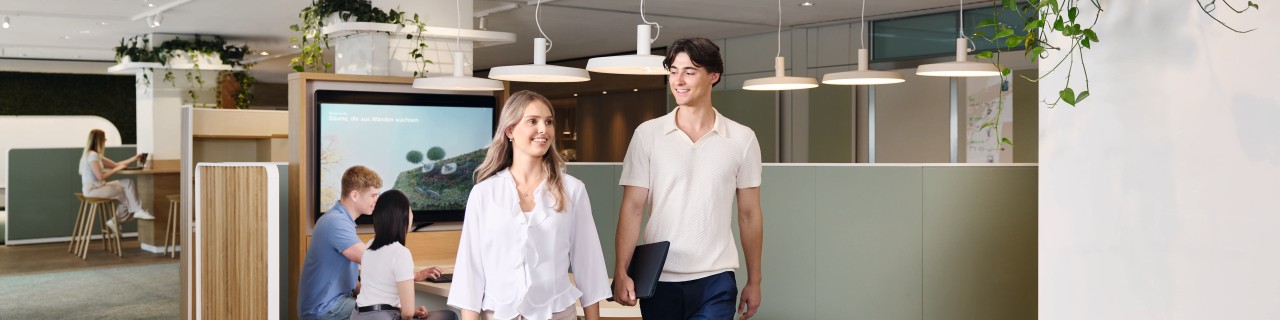 Zwei junge Nachwuchskräfte bewegen sich dynamisch von einer Gruppenarbeit weg, um zu dem nächsten Meeting zu gehen. Sie hat blonde mittellange Haare und trägt eine weiße Bluse sowie beige Hose. Er hat dunkle Haare und trägt ein helles Oberteil sowie dunkelblaue Hose und hält ein Laptop in der Hand. Im Hintergrund sitzen zwei weitere Nachwuchskräfte zusammen, die an dem Thema Verbesserung des Stadtklimas arbeiten.