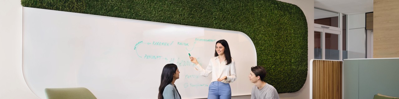 Drei junge Nachwuchskräfte befinden sich an einem Whiteboard im Digi-Café der KfW und brainstormen engagiert zusammen zum Thema hashtag Team KfW. Eine junge Frau mit dunklen mittellangen Haaren steht dabei mit einem Stift an dem Board, welches mit einer grünen dicht bewachsenen Mooswand umrandet ist. Eine weitere junge Frau mit dunklen langen Haaren sowie ein junger Mann mit braunen Haaren sitzen auf Hockern daneben und besprechen die Ergebnisse. Sie hält ein Tablet in der Hand und gleicht die Punkte mit ihren Notizen ab.