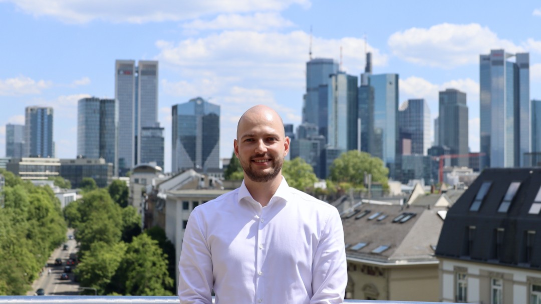 Maximillian Neeb, Mitarbeiter der KfW Konzernentwicklung trägt ein weißes Hemd und befindet sich auf der Dachterasse der KfW. Hinter ihm ist die Skyline von Frankfurt zu sehen.
