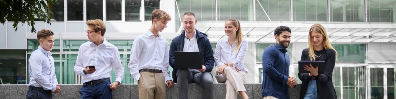 Eine diverse Gruppe von sieben Nachwuchskräften befindet sich vor einem modernen KfW Gebäude. Die Personen sind Business Casual angezogen. Manche stehen, andere sitzen auf einer niedrigen Mauer. Sie tauschen sich aus und schauen dabei auf Tablets oder Laptops.