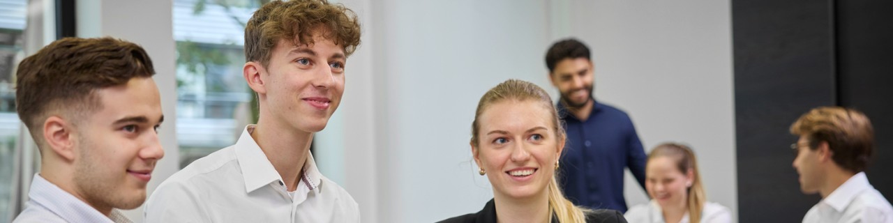 Eine diverse Gruppe von Nachwuchskräften, die an zwei Tischen verteilt ist, bespricht in kleinen Gruppen die nächsten Projekte. Die drei Personen im vorderen Teil des Bildes hören gespannt ihrem Arbeitskollegen zu.