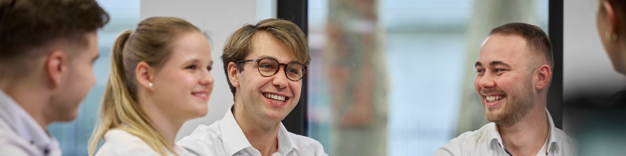 Fünf Nachwuchskräfte sitzen an einem Tisch und sind dabei in eine angeregte Diskussion verwickelt. Auf dem Tisch stehen mehrere Laptops, Tassen und Gläser.