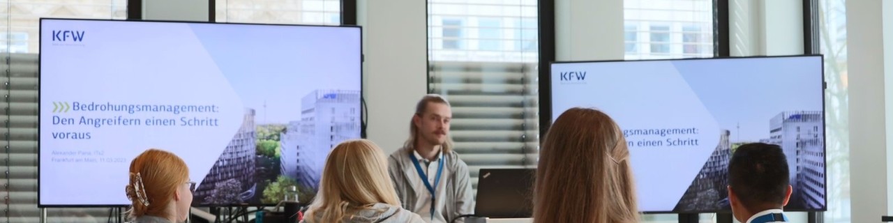 Der KfW ITler Alexander Pana sitzt vor einer Gruppe von Männern und Frauen, die an dem Hackathon Security Challenge Vortrag teilnehmen. Hinter Alexander sind zwei Bildschirme aufgebaut, auf welchen eine Präsentation mit dem Titel „Bedrohungsmanagement: Den Angreifern einen Schritt voraus“ abgespielt wird.