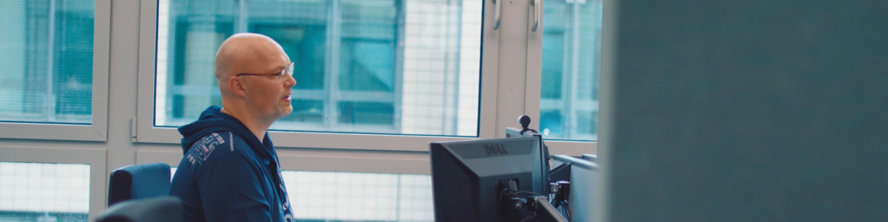 Ein Mitarbeiter aus dem Bereich IT-Security sitzt an seinem Arbeitsplatz in Berlin. Ausgestatte mit zwei Monitoren und höher verstellbarem Schreibtisch. Er trägt eine Brille, Glatze und einen blauen Kapuzen Pullover.
