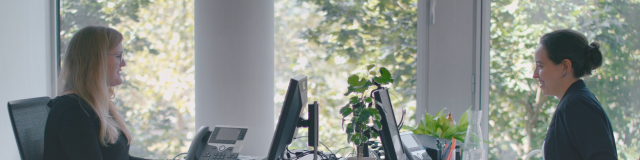 Zwei Technische Sachverständige teilen sich ein Büro mit Blick ins Grüne. Ihre Schreibtische stehen in der Mitte aneinander. Beide Frauen sitzen an ihrem Arbeitsplatz und arbeiten am Computer.