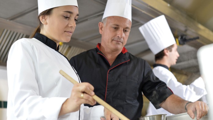 Eine Azubildende zur Restaurantfachfrau ist in der Küche und überpüft die Speisen.