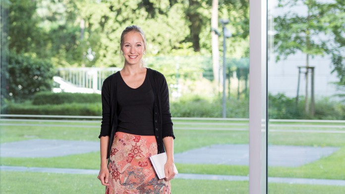 Ein Portraitfoto von Christina Wolf. Sie steht im Haupthaus vor der großen und hellen Fensterfront. Es sit Sommer und das Gras, die Bäume und Sträucher sind im satten Grün.