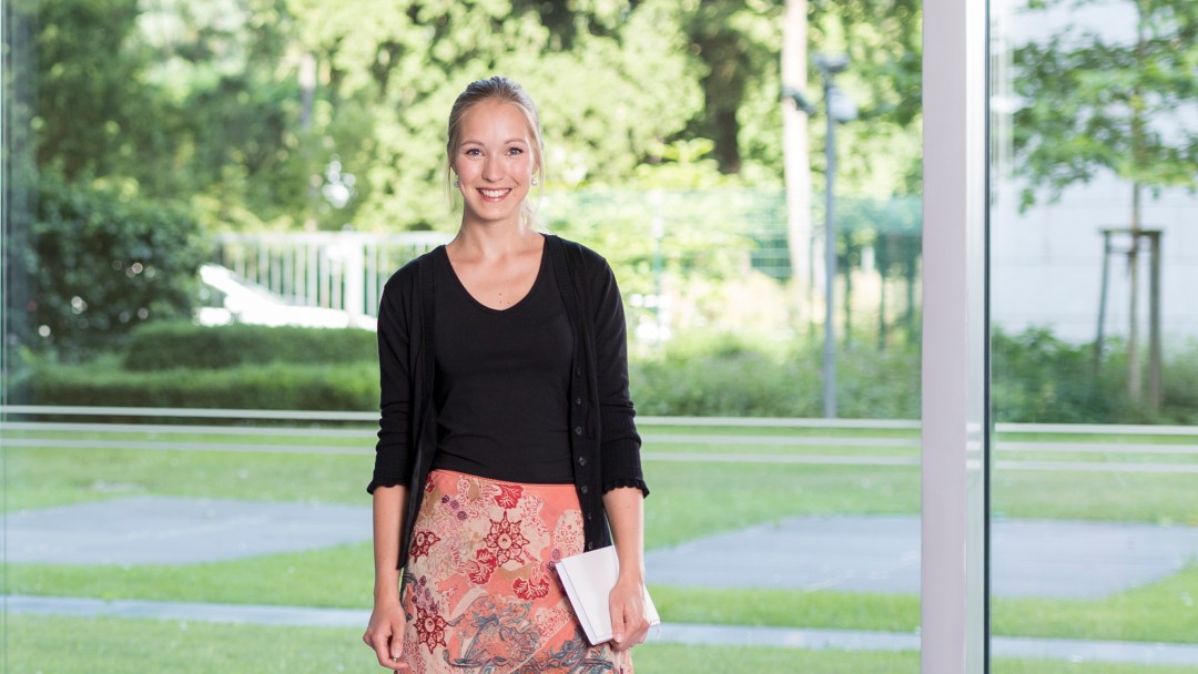 Ein Portraitfoto von Christina Wolf. Sie steht im Haupthaus vor der großen und hellen Fensterfront. Es sit Sommer und das Gras, die Bäume und Sträucher sind im satten Grün.