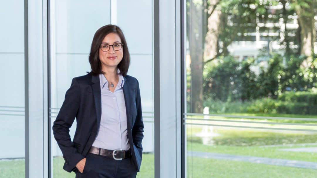 Ein Portraitfoto von Frau Dr. Lo. Sie steht im Haupthaus vor der großen Fensterfront. Draussen ist es Sommer und schönes, sonniges Wetter. 