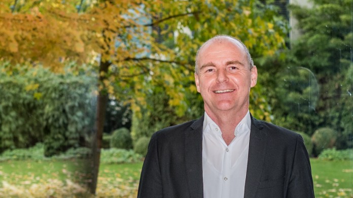 Dirk Wenzel ist technischer Sachverständiger bei der KfW. Er trägt einen schwarzen Anzug und ein weißes Hemd. Er steht im Foyer der KfW IPEX-Bank mit Blick ins Grüne.