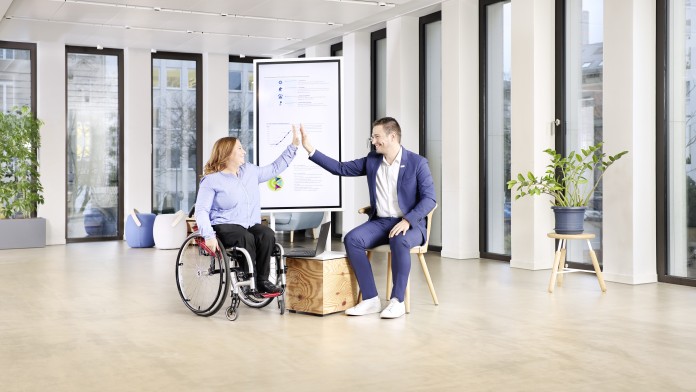 Eine Frau mit blonden Haaren und hellblauem Hemd im Rollstuhl und ein Mann mit weißem Hemd und blauen Anzug schlagen mit High Five ein, nachdem beide eine erfolgreiche Präsentation gehalten haben.