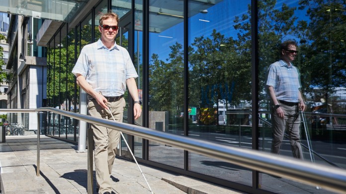 Portraitbild von Boris Spellmeier, Mitarbeiter in der KfW Entwicklungsbank. Er hat hellbraune kurze Haare, trägt eine dunkle Brille, ein kurzärmiges hellblau kariertes Hemd und eine beige Hose. Er läuft mit Unterstützung eines weißen Langstocks im Außenbereich vor einem KfW Gebäude.