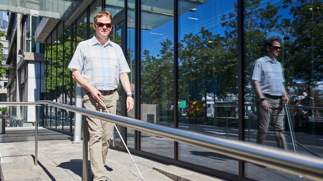 Portraitbild von Boris Spellmeier, Mitarbeiter in der KfW Entwicklungsbank. Er hat hellbraune kurze Haare, trägt eine dunkle Brille, ein kurzärmiges hellblau kariertes Hemd und eine beige Hose. Er läuft mit Unterstützung eines weißen Langstocks im Außenbereich vor einem KfW Gebäude.