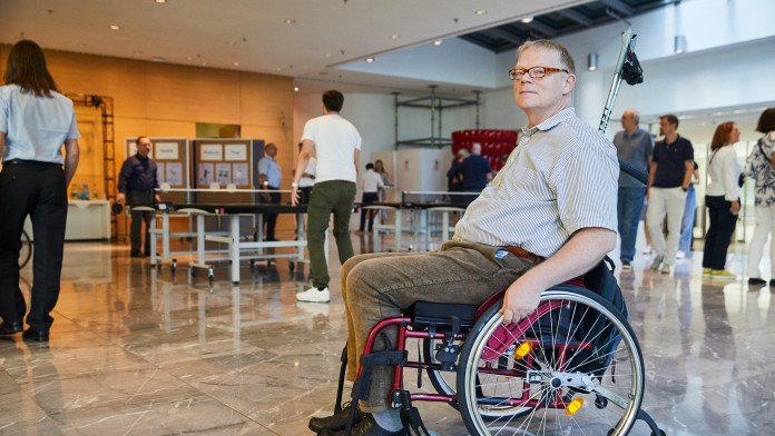 Portraitbild von Dr. Rainer Durth, Mitarbeiter in der KfW Entwicklungsbank. Er hat blonde kurze Haare, trägt eine eckige braune Brille, ein kurzärmiges gestreiftes Hemd und eine braune Kordhose. Er sitz in einem roten Rollstuhl und im Hintergrund sieht man verschiedene KfW Mitarbeitende während der KfW Gesundheitstage. 