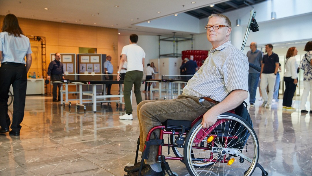 Portraitbild von Dr. Rainer Durth, Mitarbeiter in der KfW Entwicklungsbank. Er hat blonde kurze Haare, trägt eine eckige braune Brille, ein kurzärmiges gestreiftes Hemd und eine braune Kordhose. Er sitz in einem roten Rollstuhl und im Hintergrund sieht man verschiedene KfW Mitarbeitende während der KfW Inklusions- und Gesundheitstage. 