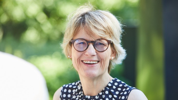 Portraitbild von Esther Bach, Mitarbeiterin in der KfW Entwicklungsbank. Sie hat blonde kurze Haare, trägt eine braune Brille und ein dunkles, weiß gepunktetes kurzärmiges Oberteil.