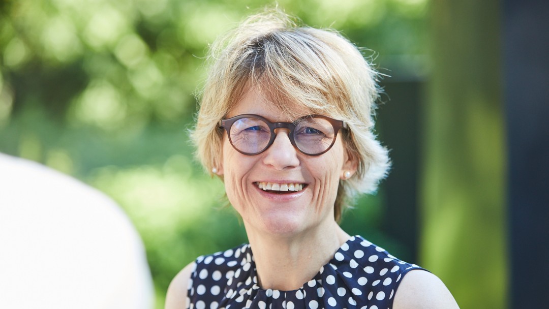 Portraitbild von Esther Bach, Mitarbeiterin in der KfW Entwicklungsbank. Sie hat blonde kurze Haare, trägt eine braune Brille und ein dunkles, weiß gepunktetes kurzärmiges Oberteil.